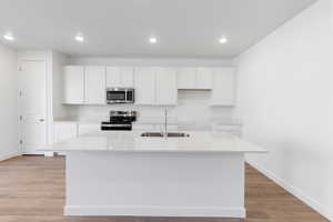 Kitchen with white cabinets, appliances with stainless steel finishes, light wood-style floors, light stone countertops, and an island with sink