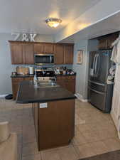 Kitchen with a textured ceiling, stainless steel appliances, dark countertops, light tile patterned flooring, and an island with sink