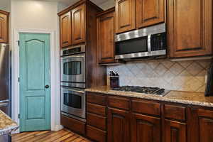Kitchen with stainless steel appliances, light stone countertops, light wood-style floors, tasteful backsplash, and brown cabinets