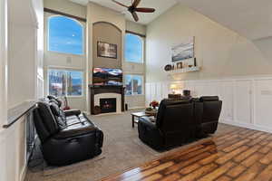 Carpeted living area featuring a decorative wall, a towering ceiling, a tiled fireplace, wainscoting, and a ceiling fan