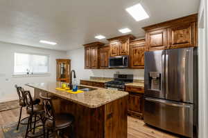 Kitchen with stainless steel appliances, light wood finished floors, an island with sink, light stone countertops, and a breakfast bar area
