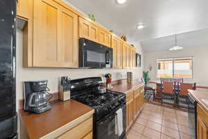 Kitchen with black appliances, decorative light fixtures, light tile patterned floors, light brown cabinets, and lofted ceiling