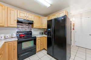 Kitchen featuring black appliances, light countertops, backsplash, light tile patterned floors, and under cabinet range hood