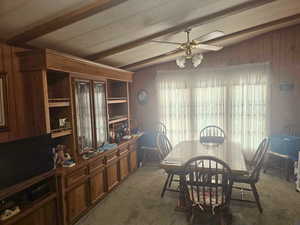 Dining area with wood walls, dark colored carpet, ceiling fan, and beamed ceiling