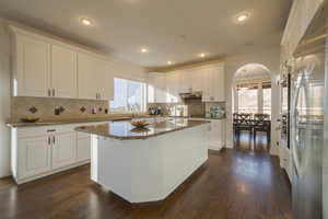 Kitchen with dark stone counters, arched walkways, white cabinets, stainless steel fridge, and dark wood flooring