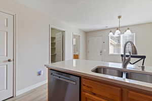 Kitchen with stainless steel dishwasher, light stone countertops, light wood-type flooring, brown cabinets, and decorative light fixtures