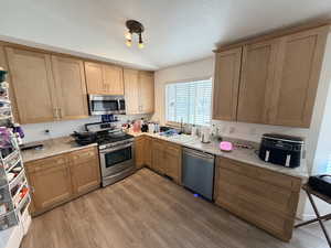 Kitchen featuring appliances with stainless steel finishes, light wood-style floors, lofted ceiling, light stone counters, and brown cabinetry