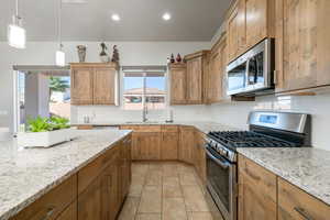 Kitchen with stainless steel appliances, brown cabinetry, decorative light fixtures, light stone countertops, and recessed lighting
