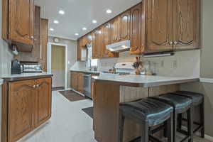 Kitchen with a breakfast bar area, recessed lighting, brown cabinets, white range, and under cabinet range hood