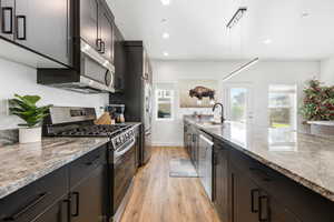 Kitchen featuring appliances with stainless steel finishes, light stone counters, light wood-type flooring, recessed lighting, and dark cabinets