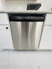 Kitchen view of stainless steel dishwasher, white cabinetry, and light stone countertops