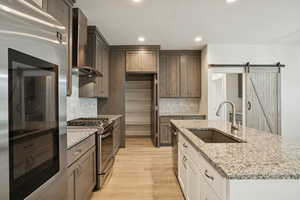 Kitchen featuring a barn door, stainless steel appliances, light stone countertops, wall chimney range hood, and white cabinetry