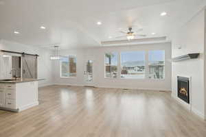 Unfurnished living room with a barn door, a tray ceiling, recessed lighting, light wood-type flooring, and a glass covered fireplace