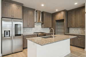 Kitchen with stainless steel appliances, light stone counters, wall chimney range hood, backsplash, and light wood-style floors
