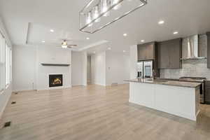 Kitchen with an island with sink, wall chimney range hood, appliances with stainless steel finishes, tasteful backsplash, and light stone counters