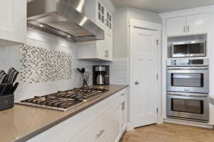 Kitchen with wall chimney exhaust hood, stainless steel appliances, white cabinetry, light wood-type flooring, and tasteful backsplash