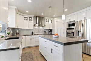 Kitchen featuring a kitchen island, stainless steel appliances, wall chimney range hood, white cabinetry, and backsplash