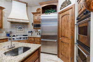 Kitchen with brown cabinets, stainless steel appliances, premium range hood, backsplash, and light stone countertops