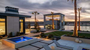 Back of property at dusk featuring a water view, a patio, and an in-ground hot tub