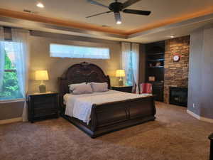 Carpeted bedroom featuring a fireplace, ceiling fan, and a raised ceiling