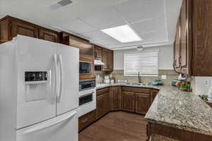 Kitchen featuring white appliances, a drop ceiling, dark wood-type flooring, dark brown cabinets, and backsplash