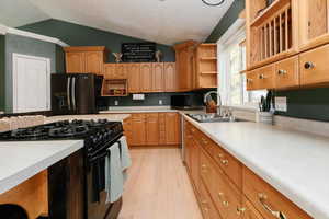 Kitchen featuring open shelves, black appliances, vaulted ceiling, light countertops, and light wood-style floors