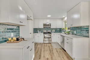 Kitchen with appliances with stainless steel finishes, white cabinets, light wood finished floors, decorative backsplash, and light stone countertops
