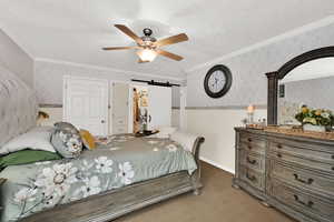 Carpeted bedroom featuring wallpapered walls, a barn door, ornamental molding, ceiling fan, and a textured ceiling
