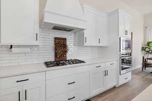 Kitchen with white cabinetry, custom exhaust hood, light wood-type flooring, stainless steel appliances, and light stone countertops