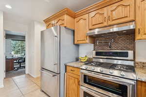 Kitchen featuring appliances with stainless steel finishes, under cabinet range hood, light stone counters, light tile patterned flooring, and a desk