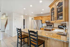 Kitchen with a breakfast bar, recessed lighting, appliances with stainless steel finishes, light stone counters, and glass insert cabinets