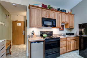 Kitchen with black appliances, light tile patterned floors, vaulted ceiling, and light stone counters
