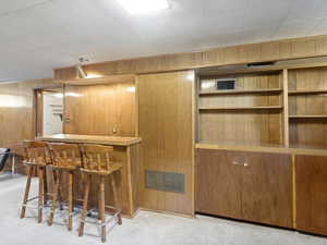 Indoor dry bar with wood walls, light carpet, brown cabinetry, light countertops, and open shelves