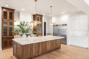 Kitchen with dual tone cabinetry, stainless steel built in refrigerator, a kitchen island with sink, backsplash, and light stone countertops
