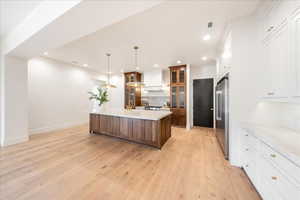 Kitchen featuring glass fronted cabinets, a kitchen island with sink, hanging light fixtures, two tone color scheme, and light wood-style floors