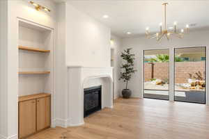 Unfurnished living room featuring built in shelves, light wood-style flooring, a chandelier, and a glass covered fireplace