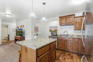 Kitchen with brown cabinetry, stainless steel appliances, light stone countertops, a kitchen island, and decorative light fixtures
