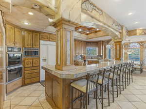 Kitchen featuring light tile patterned floors, a kitchen bar, built in appliances, brown cabinetry, and recessed lighting