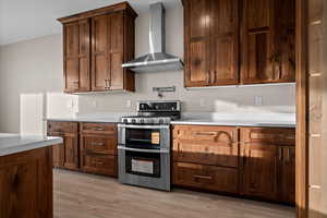 Kitchen with wall chimney range hood, double oven range, light wood-type flooring, and light stone countertops