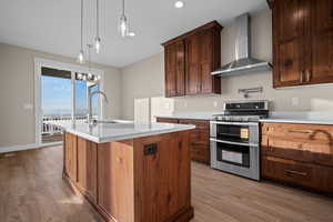 Kitchen with range with two ovens, wall chimney exhaust hood, an island with sink, hanging light fixtures, and light wood-style flooring