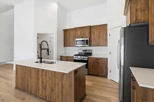 Kitchen featuring a high ceiling, stainless steel appliances, an island with sink, light wood finished floors, and brown cabinetry
