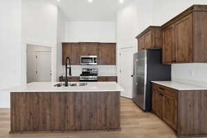Kitchen with a towering ceiling, stainless steel appliances, a kitchen island with sink, light wood-style floors, and light stone countertops