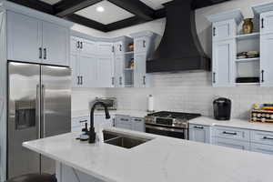 Kitchen with open shelves, premium range hood, stainless steel appliances, light stone counters, and beamed ceiling