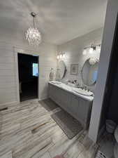 Full bath with double vanity, a textured ceiling, a chandelier, light wood-type flooring, and wooden walls