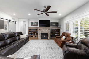 Living room with a ceiling fan, a stone fireplace, a barn door, recessed lighting, and wood finished floors.