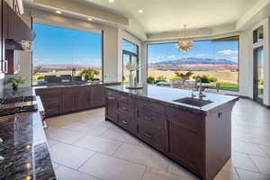 Kitchen with dark stone countertops, hanging light fixtures, an island with sink, under cabinet range hood, and dark brown cabinets