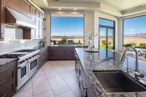Kitchen with range with two ovens, dark stone counters, light tile patterned floors, tasteful backsplash, and range hood