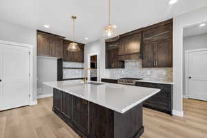Kitchen with tasteful backsplash, dark brown cabinetry, light stone countertops, a center island with sink, and recessed lighting