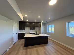 Kitchen featuring dark brown cabinetry, a kitchen island with sink, recessed lighting, light wood-style floors, and light stone counters
