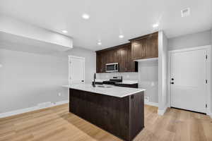 Kitchen with dark brown cabinetry, a kitchen island with sink, stainless steel appliances, light wood-style flooring, and light stone countertops